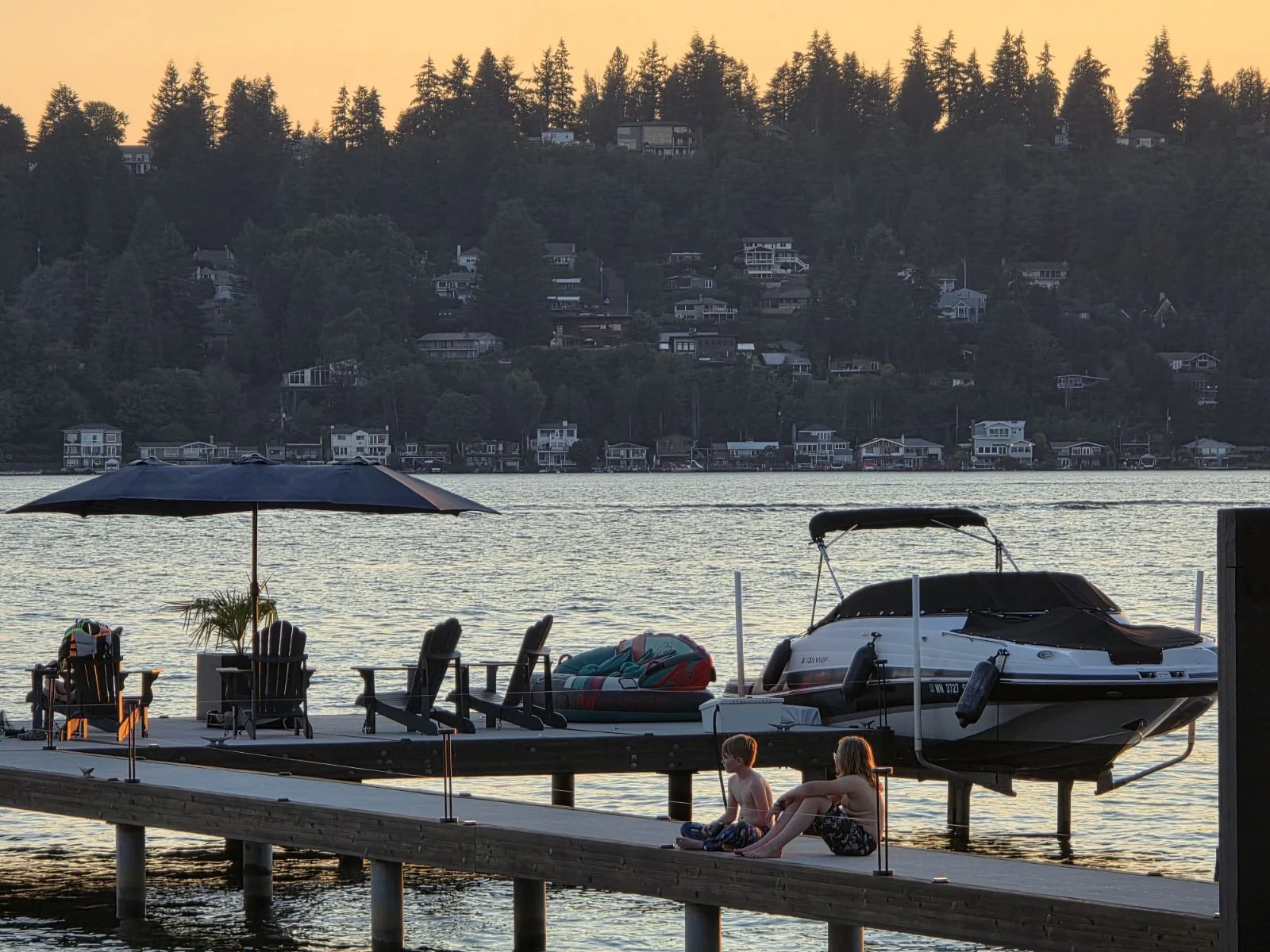 Children sitting on a dock by the water at sunset near boats.